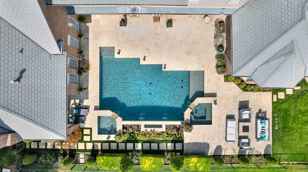 A straight-down aerial shot highlights the geometric luxury of the swimming pool, dual integrated hot tub, and surrounding travertine decking. Built-in fire features, sunbathing ledges, and landscaped pathways show off the backyard's layout.