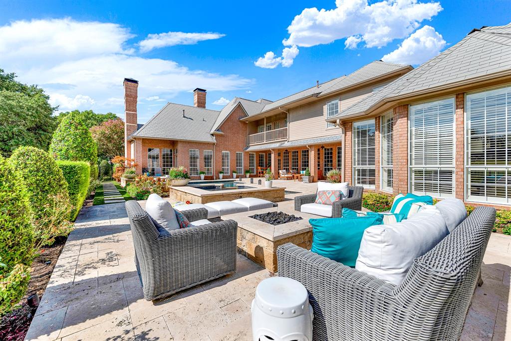 Travertine pavers create a sprawling rear patio outfitted with an integrated fire pit and cozy wicker lounge seating. The classic brick-and-siding exterior frames this private outdoor sanctuary, complete with views of the pool and lush perimeter landscaping.