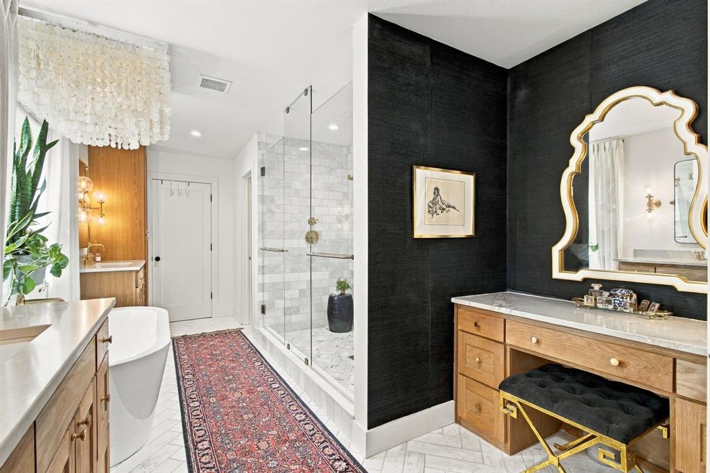 Glass-enclosed shower with subway tile surround, standalone soaking tub, dual vanities with integrated sinks, wood-finish cabinetry, and a dedicated vanity desk with a stone countertop