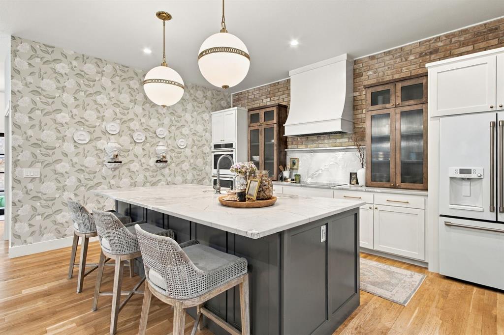 Spacious kitchen island with a light-toned countertop, wood-finish flooring, two spherical pendant lights, exposed brick accent wall, and integrated stainless steel appliances