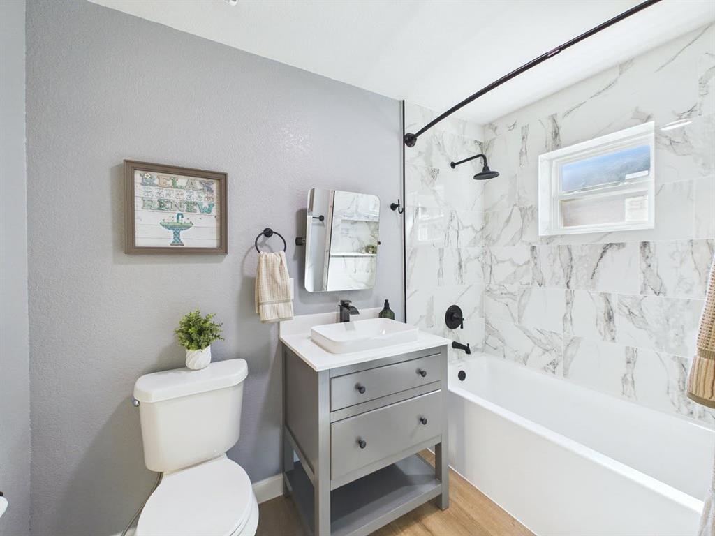 Wall-mounted shower head with matte black fixtures, white rectangular bathtub, wood-finish flooring, vanity with integrated vessel sink, and large format wall tile