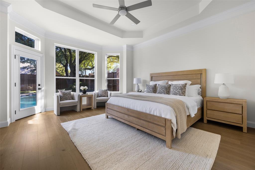 Spacious first-floor primary suite features a tray ceiling, wide-plank wood flooring, contemporary ceiling fan, sitting area, and expansive windows overlooking the backyard and pool.