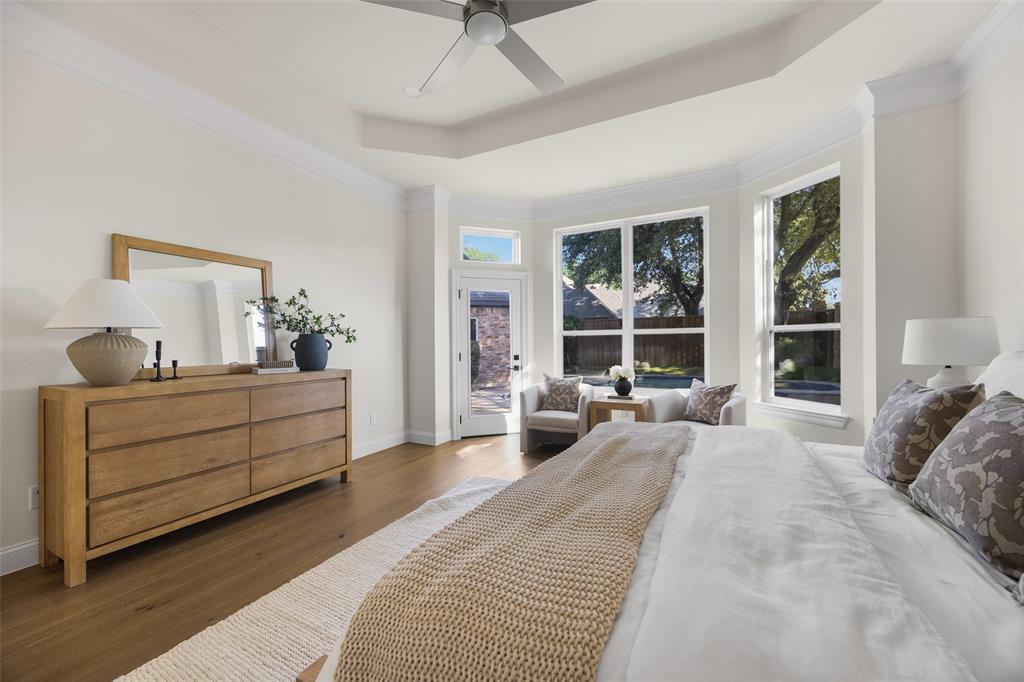 Spacious first-floor primary suite features a tray ceiling, wide-plank wood flooring, contemporary ceiling fan, sitting area, and expansive windows overlooking the backyard and pool.