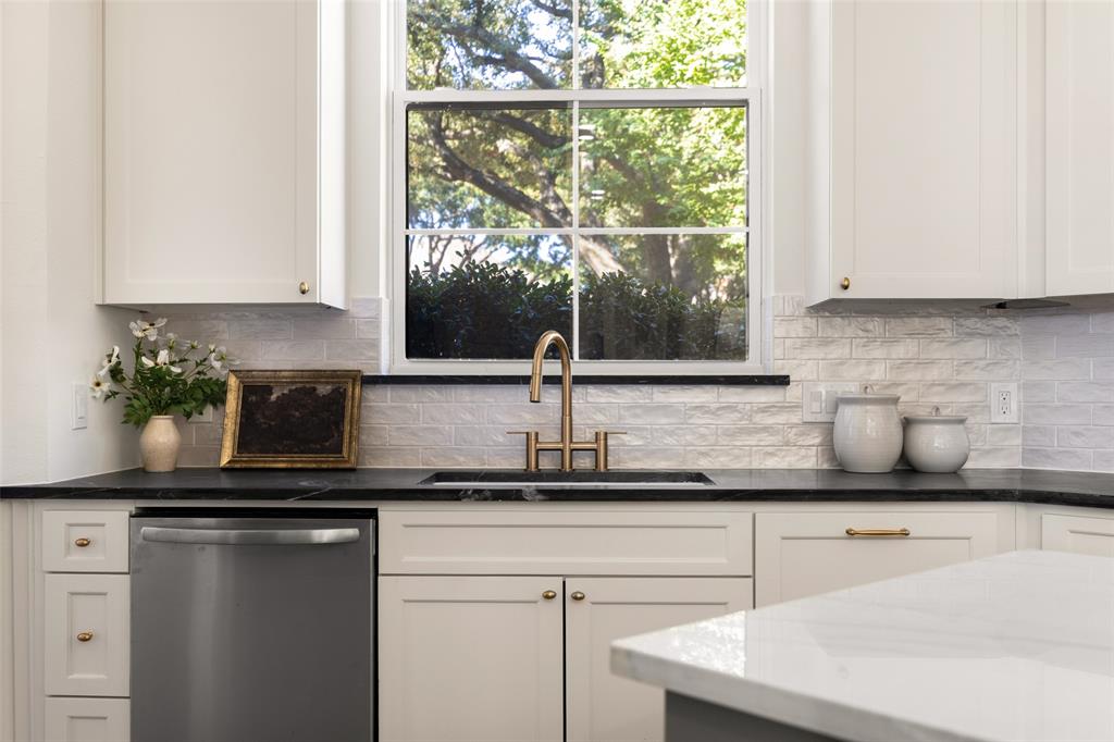 Oversized kitchen window fills the workspace with natural light, highlighting the dark perimeter countertops, undermount sink, brass-toned faucet, stainless-steel dishwasher, and crisp cabinetry.