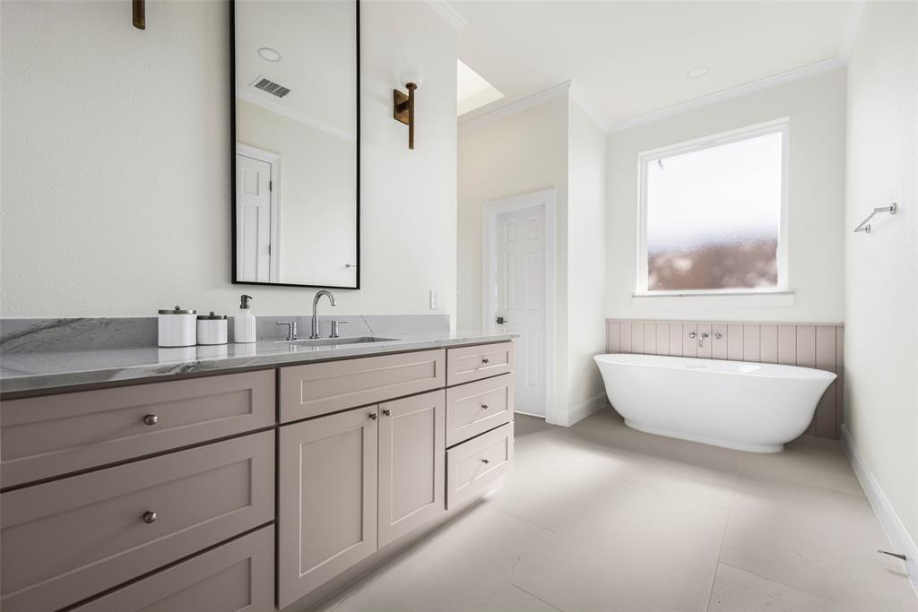 Serene primary bath featuring a freestanding soaking tub beneath a large window, separate vanity, large-format tile flooring, tailored cabinetry, and understated contemporary finishes.