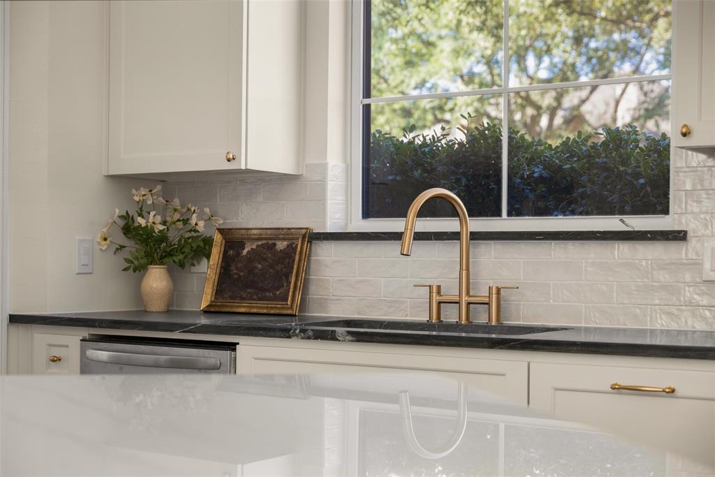 Thoughtfully selected finishes include dark perimeter countertops, a brass-toned bridge faucet, textured white backsplash tile, stainless-steel appliances, and cabinetry extending around the window for additional storage.