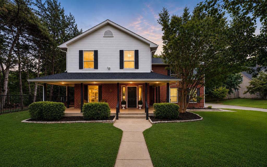 Picture-perfect curb appeal with a charming wraparound-style covered porch, mature trees, manicured landscaping, and a timeless brick-and-siding exterior.