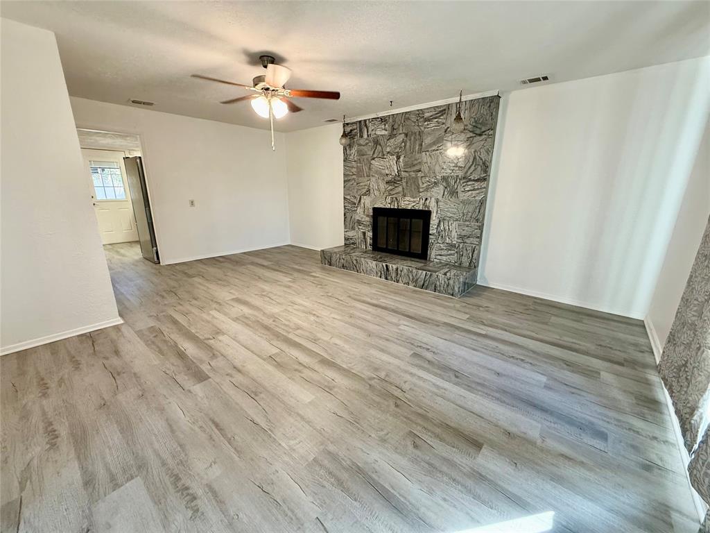 Spacious room featuring wood-finish flooring, a stone fireplace with hearth, and a ceiling fan with light fixture Spacious room featuring wood-finish flooring, a stone fireplace with hearth, and a ceiling fan with light fixture