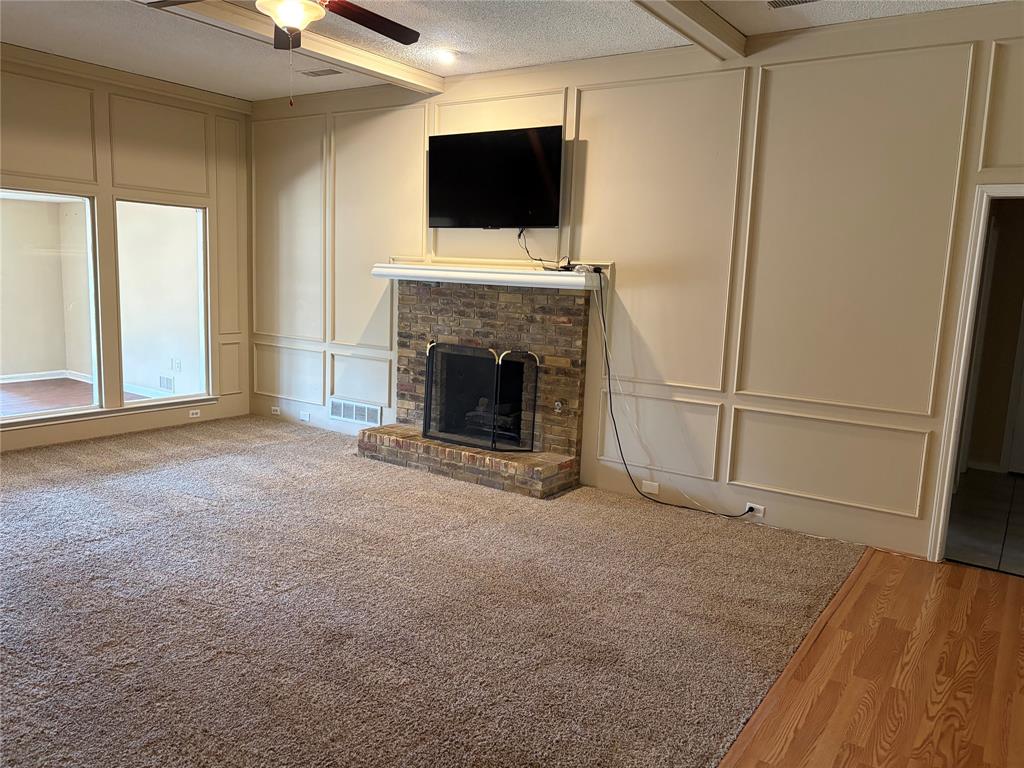 Stone-faced fireplace with a white mantel, large picture windows, decorative wall paneling, ceiling fan with light fixture, and wood-finish flooring