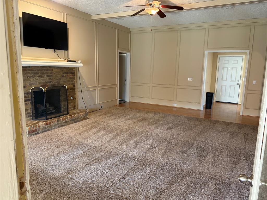 Living area featuring a brick fireplace with a white mantel, decorative wall paneling, a ceiling fan with integrated lighting, and a mix of carpeted and wood-finish flooring