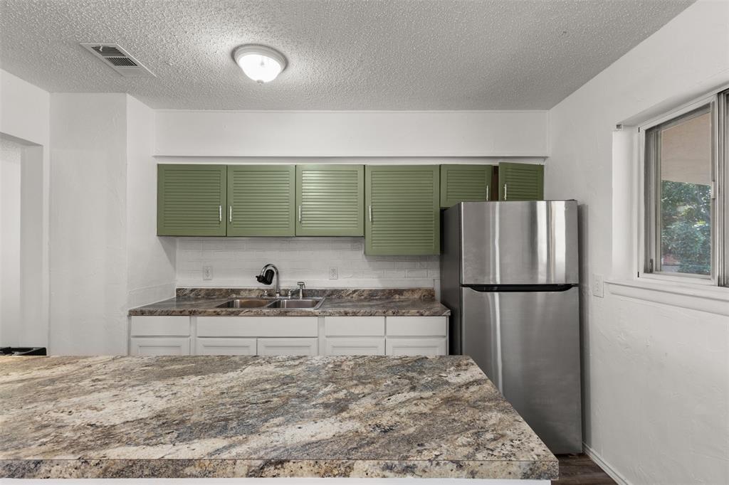 Kitchen featuring stone countertops, a double basin sink, green louvered upper cabinetry, white lower cabinetry, and a stainless steel refrigerator