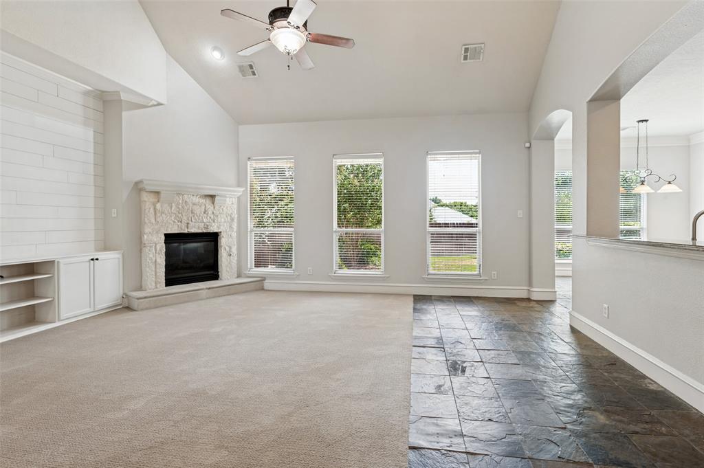 Vaulted ceiling with a ceiling fan, stone-clad fireplace, built-in cabinetry, large windows, and a mix of carpet and stone tile flooring