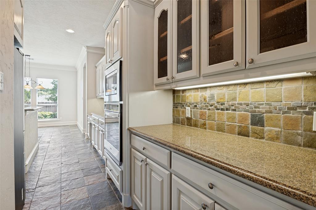 White cabinetry with glass-front upper cabinets, light-toned stone tile flooring, built-in stainless steel oven, stone tile backsplash, and granite-finish countertops