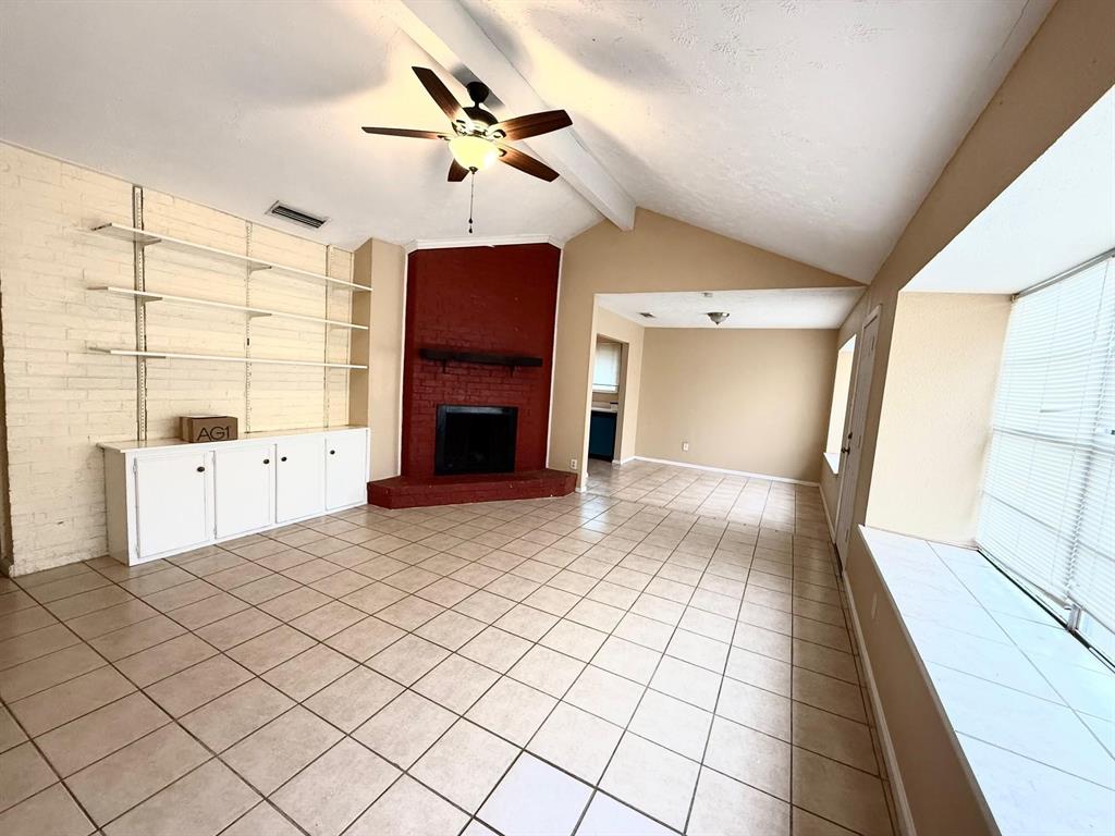 Vaulted ceiling with a ceiling fan, tiled flooring, brick accent wall with built-in shelving and cabinetry, and a brick fireplace