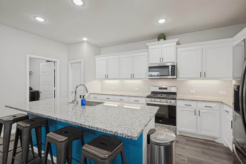 Kitchen featuring a central island with a sink and seating, white cabinetry, stainless steel appliances, granite countertops, and wood-finish flooring Kitchen featuring a central island with a sink and seating, white cabinetry, stainless steel appliances, granite countertops, and wood-finish flooring