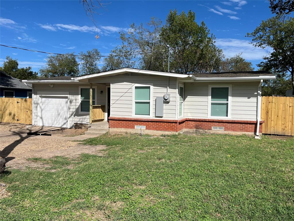 Single-story residence featuring light-toned horizontal siding, a red brick masonry base, an attached single-car garage, and wood privacy fencing Single-story residence featuring light-toned horizontal siding, a red brick masonry base, an attached single-car garage, and wood privacy fencing