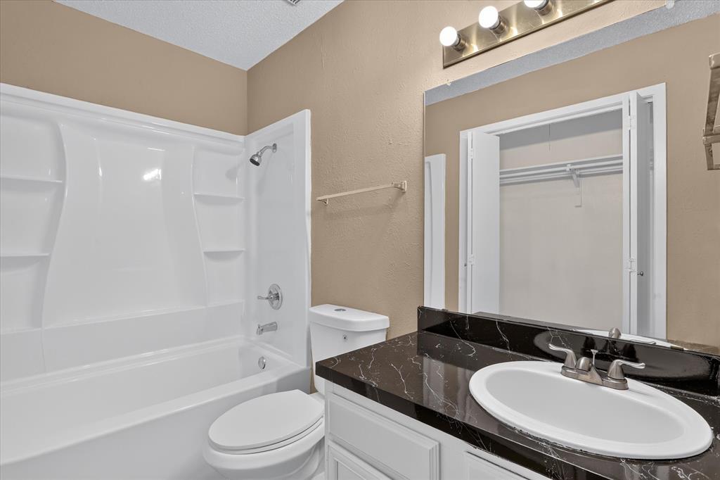 White bathtub with integrated shower, single vanity with oval sink, dark countertop with light veining, and wall-mounted mirror