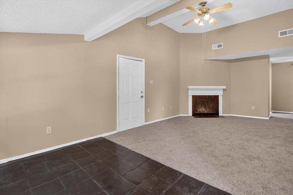 Vaulted ceiling with exposed beams, ceiling fan light fixture, white mantel over fireplace, mixed flooring with carpet and dark tile, and paneled interior door