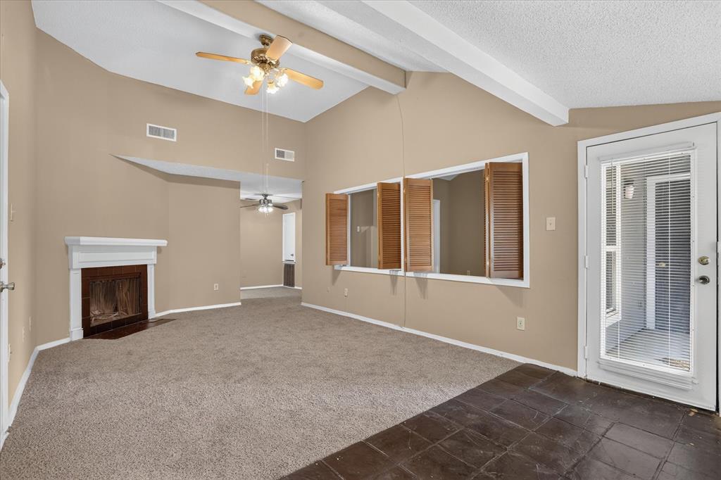 Vaulted ceiling with exposed beams, ceiling fan, fireplace with white mantel, windows with wooden shutters, and a glass-paneled exterior door