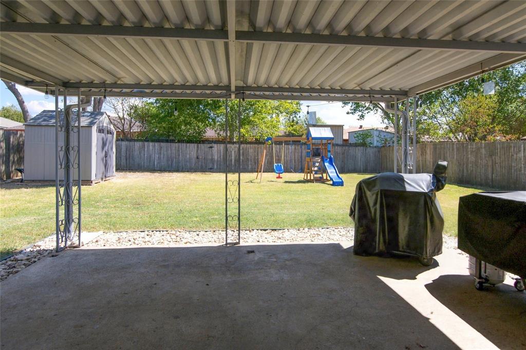 Covered concrete patio with decorative metal supports, overlooking a grassy yard with a privacy fence and a storage shed