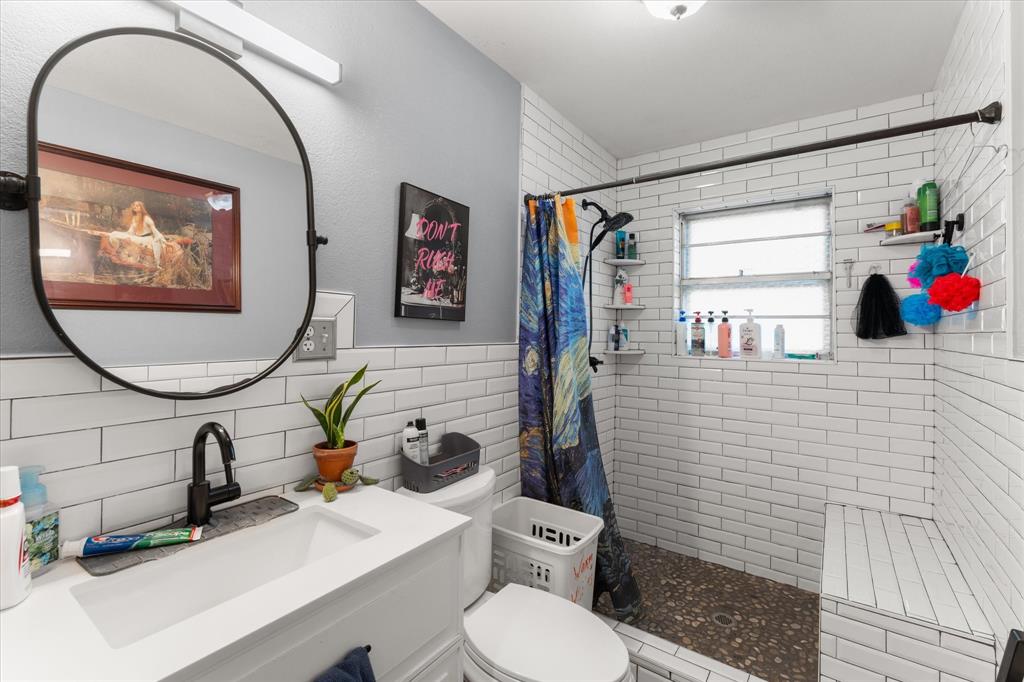 Fully remodeled Bathroom featuring a white vanity with an integrated rectangular sink, matte black faucet, oval wall-mounted mirror, white subway tile walls, and a walk-in shower with stone flooring