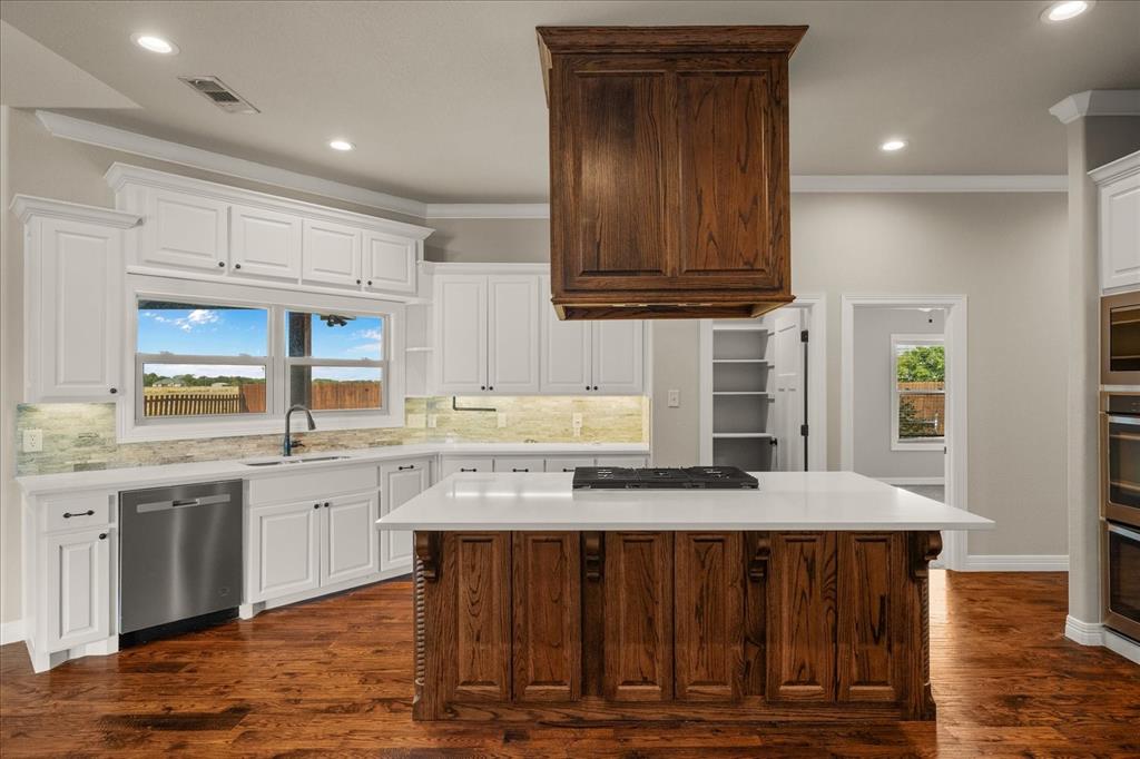 Gourmet kitchen with wood-finish flooring, white cabinetry, a large center island with a cooktop, and stainless steel appliances including a built-in oven and dishwasher