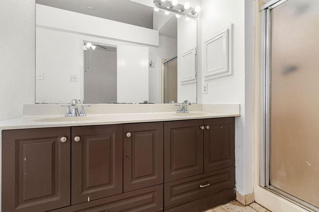 Dual-sink vanity with cabinetry, expansive wall-mounted mirror, overhead vanity lighting, and a glass-enclosed shower stall
