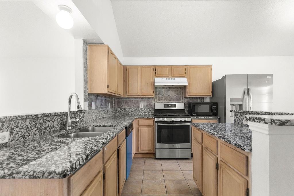 Kitchen featuring wood cabinetry, stone-finish countertops, tile flooring, stainless steel appliances, and a vaulted ceiling
