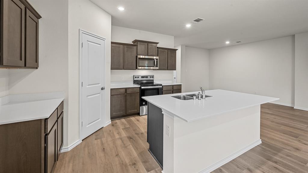 Kitchen featuring dark wood-finish cabinetry, light countertops, a kitchen island with an undermount dual-basin sink, stainless steel appliances, and recessed lighting