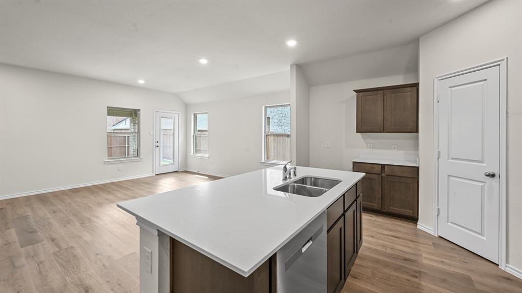 Open-concept kitchen and living area featuring a large island with white quartz countertop and integrated sink, dark wood cabinetry, stainless steel appliances, recessed ceiling lighting, wood-finish flooring throughout, and abundant natural light from multiple windows
