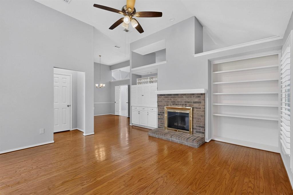 Vaulted ceiling with a ceiling fan, wood-finish flooring, brick fireplace with a brass-tone insert, built-in shelving, and built-in white cabinetry