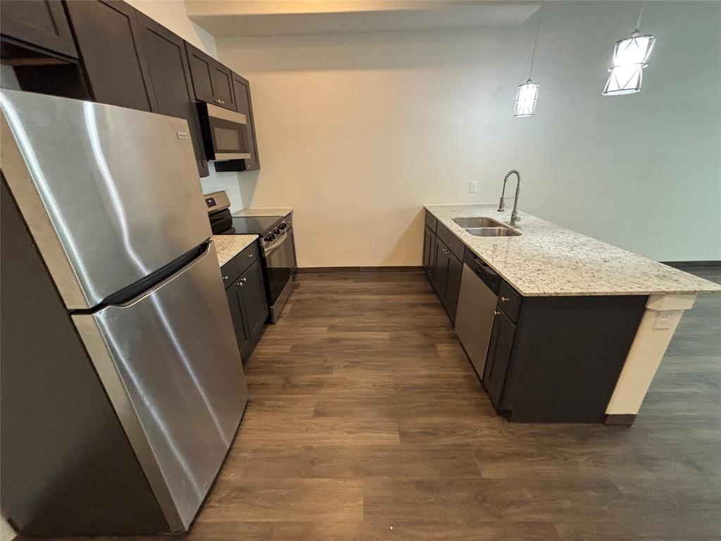 Kitchen featuring wood-finish flooring, dark cabinetry, stainless steel appliances, and a central island with a double basin sink and stone-finish countertop