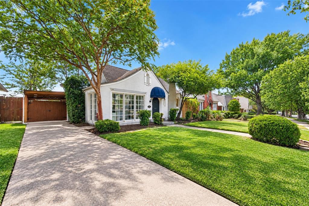White stucco exterior with a blue awning, expansive front lawn, mature trees, and a concrete driveway leading to a covered carport