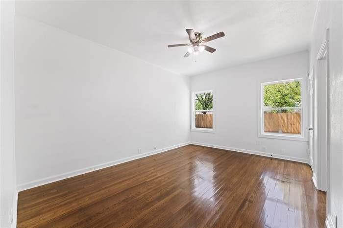 Wood-finish flooring, two windows with white trim, a ceiling fan with integrated lighting, and a white interior door
