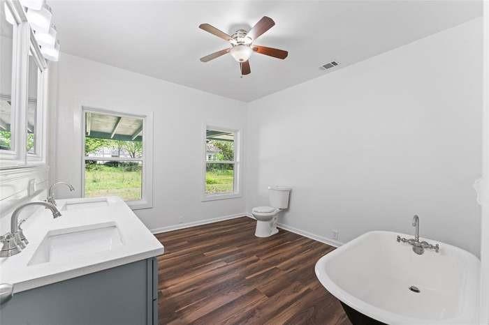 Freestanding soaking tub, wood-finish flooring, dual basin vanity with white countertop, ceiling fan with integrated lighting, and multiple windows