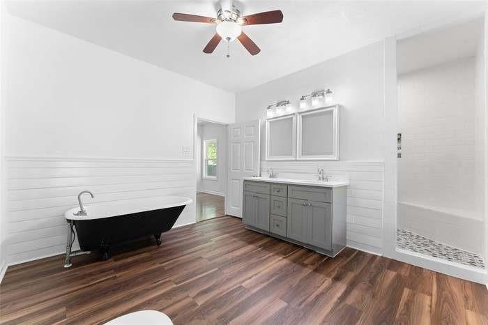 Wood-finish flooring, dual vanity with gray cabinetry, white shiplap wall treatment, freestanding clawfoot tub, and a tiled shower with a built-in bench