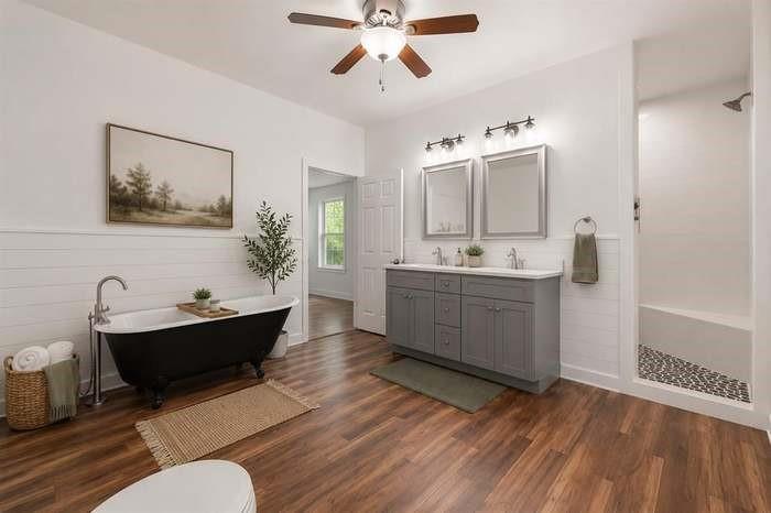Freestanding black clawfoot tub, dual vanity with shaker-style cabinetry, walk-in shower with river rock tile, wood-finish flooring, and a ceiling fan