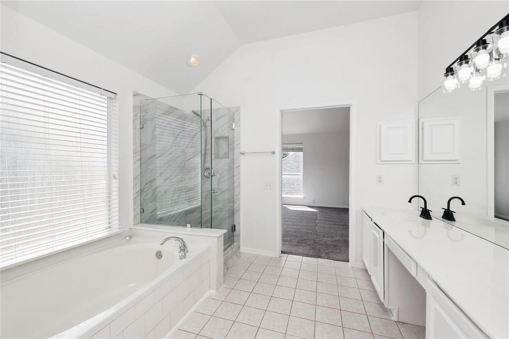 Soaking tub, glass-enclosed shower with tiled walls, dual vanity with black fixtures, vaulted ceiling, and tiled flooring