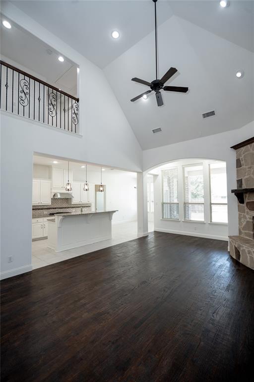 Vaulted ceiling with a ceiling fan, dark wood-finish flooring, stone fireplace, and an open-concept kitchen with a breakfast bar