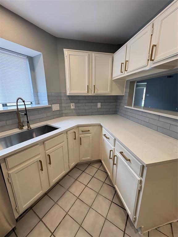 White cabinetry with gold-tone hardware, light-toned countertops, subway tile backsplash, stainless steel sink, and gray tiled flooring White cabinetry with gold-tone hardware, light-toned countertops, subway tile backsplash, stainless steel sink, and gray tiled flooring