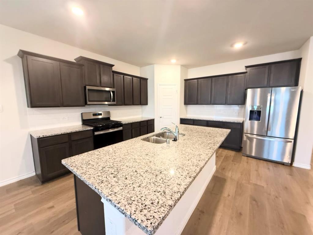 Kitchen featuring dark wood cabinetry, stone-finish countertops, stainless steel appliances, a central island with an integrated sink, and wood-finish flooring