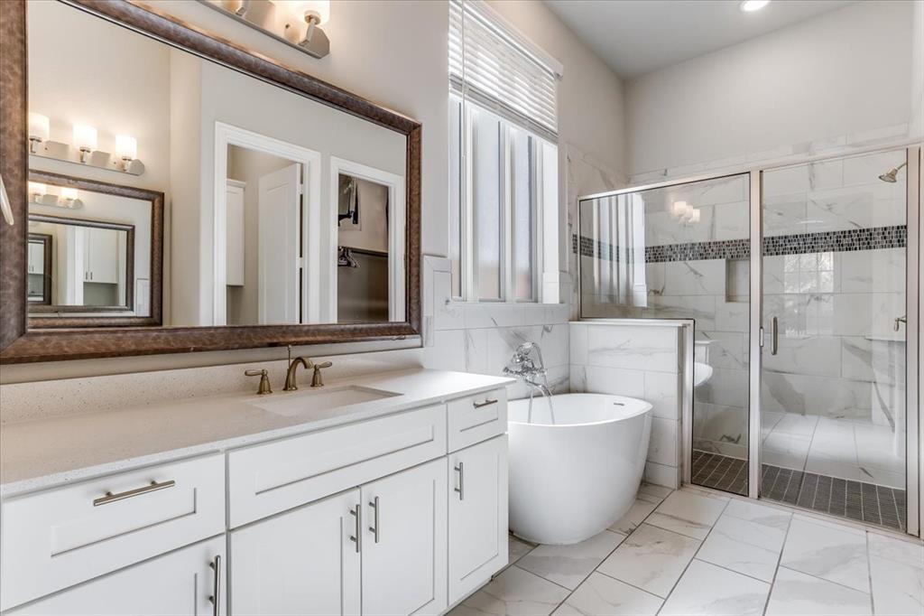 Dual-sink vanity with white cabinetry and light-toned countertop, freestanding soaking tub, glass-enclosed shower with tiled walls, and large framed wall mirror