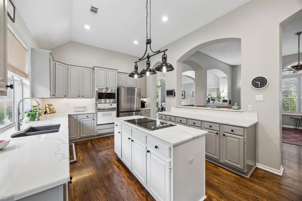 Vaulted ceiling kitchen featuring an island cooktop, built-in stainless steel appliances, light-toned cabinetry, wood-finish flooring, and white countertops