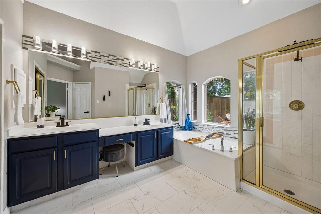 Vaulted ceiling over a bathroom featuring a large built-in tub, a glass-enclosed shower, dual vanities with dark cabinetry, and white tile flooring