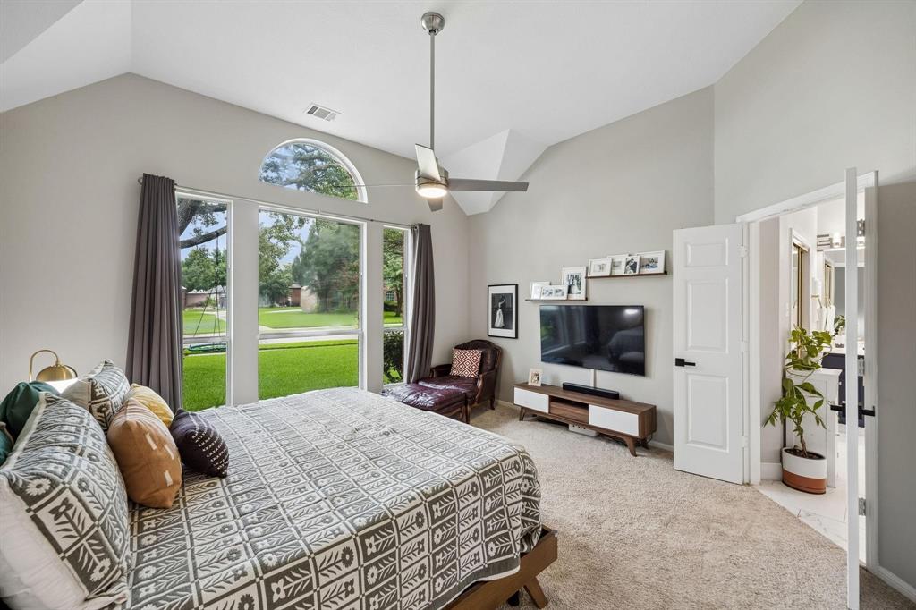 Vaulted ceiling with a ceiling fan, arched transom window, large windows with exterior landscaping, carpet flooring, and a paneled door
