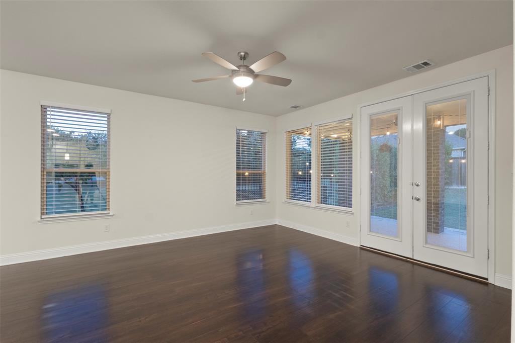 Dark wood-finish flooring, white walls, multiple windows with blinds, double glass-paneled doors, and a ceiling fan with integrated lighting