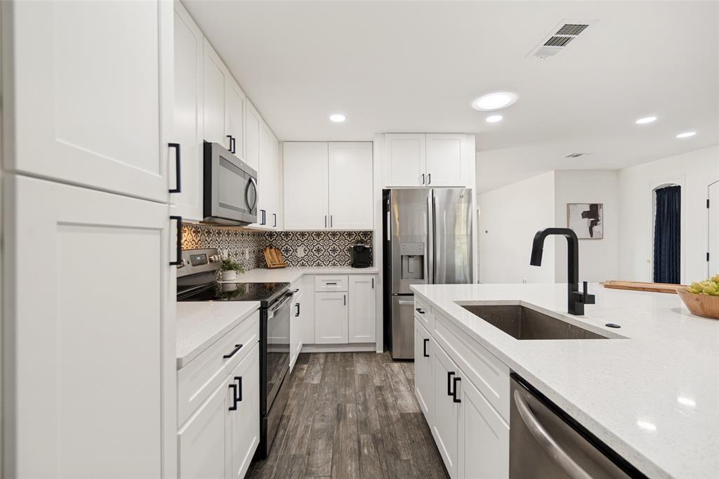 Modern kitchen featuring white shaker cabinetry, stainless steel appliances, wood-finish flooring, and a large center island with an undermount sink and matte black faucet
