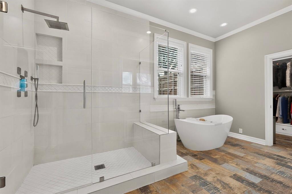 Walk-in glass-enclosed shower featuring a rainfall showerhead, handheld sprayer, and built-in tiled shelving, accompanied by a freestanding soaking tub, wood-finish flooring, and recessed lighting