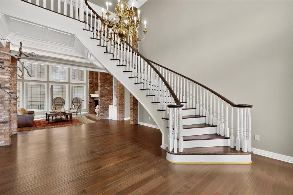 Curved staircase featuring white balusters, a dark wood handrail, and wood-finish treads, complemented by wide-plank wood-finish flooring, exposed brick structural pillars, and a multi-tiered metal chandelier
