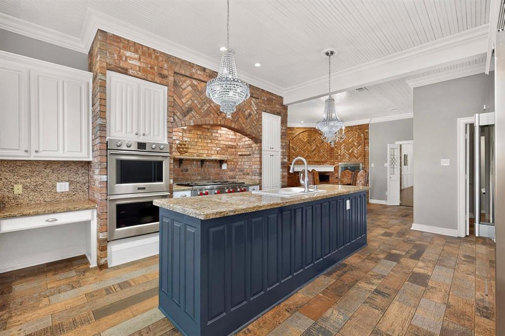 Kitchen featuring a blue-painted center island with a stone countertop, exposed brick wall with a built-in cooktop, stainless steel double wall ovens, beaded-board ceiling, and multi-toned wood-finish flooring
